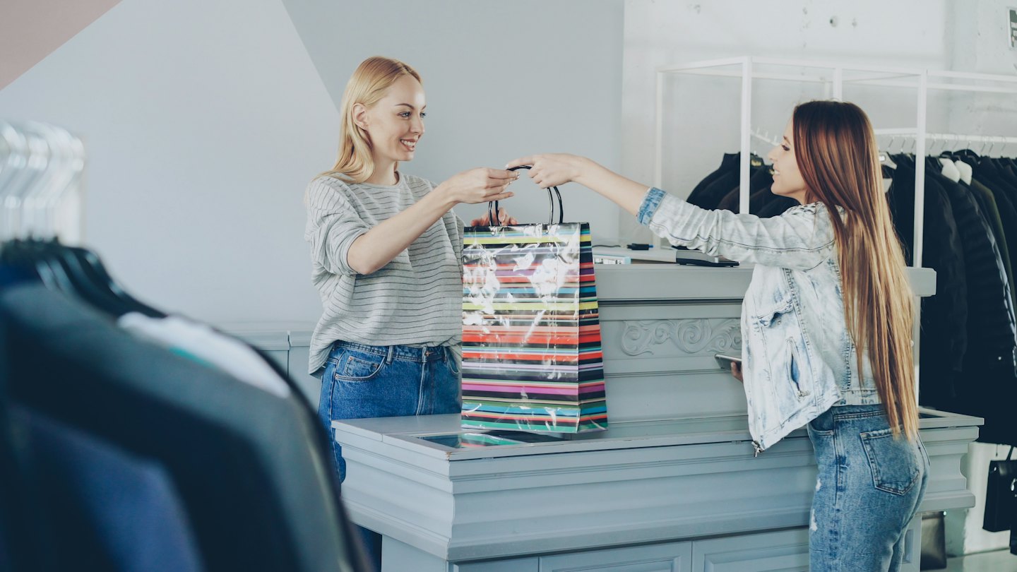 Shop assistant hands customer a colorful shopping bag at a clothing store.