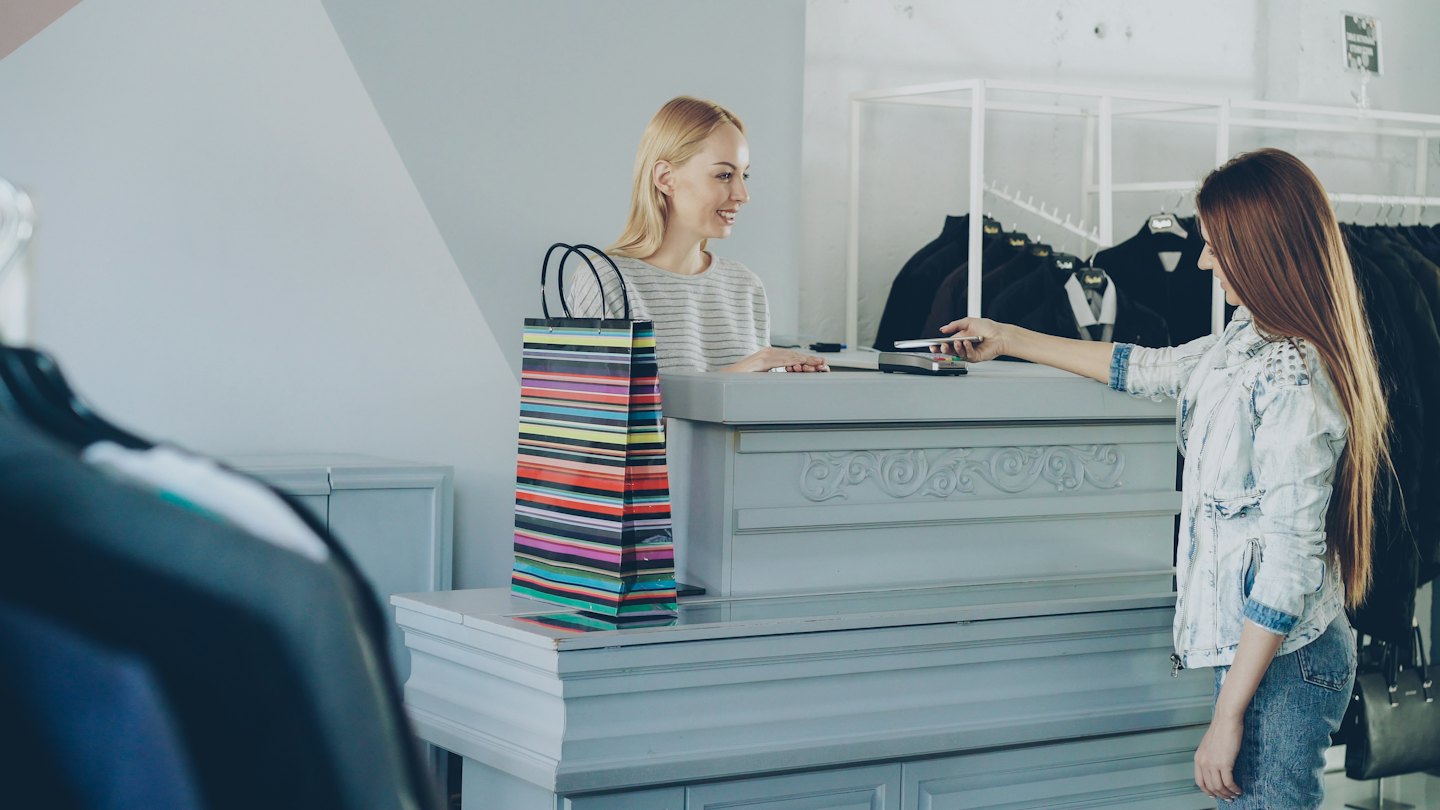 Cashier smiles as customer pays with smartphone at clothing store checkout.